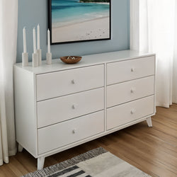 Modern white 6-drawer bedroom dresser with round knobs and mid-century legs on hardwood floor, styled with coastal wall art, candles and white curtains