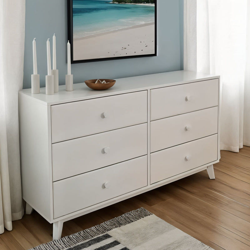Modern white 6-drawer bedroom dresser with round knobs and mid-century legs on hardwood floor, styled with coastal wall art, candles and white curtains