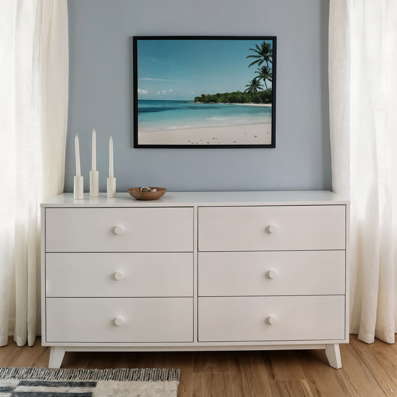 White six-drawer dresser with round knobs in minimal coastal bedroom decor, framed beach wall art, three white candles, wooden bowl, sheer curtains, hardwood floor