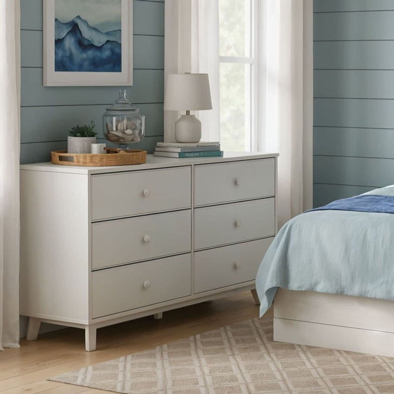White six-drawer dresser in coastal-style bedroom with blue shiplap wall, light-blue bedding, lamp, tray of shells and books, framed blue artwork and hardwood floor