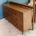 Solid wood six-drawer mid-century modern dresser with natural finish beside baby crib in blue nursery on light hardwood floor
