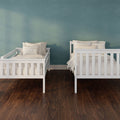 Two white wooden twin beds with neutral striped pillows in a modern nursery against a teal textured wall and dark hardwood floor