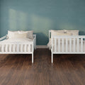 Two white wooden twin beds with beige pillows and striped lumbar cushions against a teal textured accent wall in a minimalist children's shared bedroom with dark hardwood floors