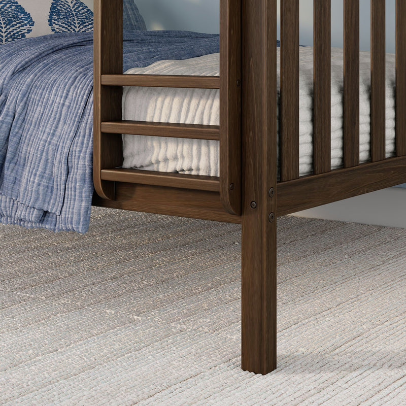 Close-up of walnut-finish wooden crib rail with blue quilted blanket and white ribbed mattress pad on textured neutral area rug — nursery bedding detail