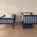 Two navy blue twin beds with white and floral bedding in a kids’ bedroom, light hardwood floors, neutral beige wall and under-bed storage drawer