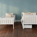 Nursery with white wooden twin beds, neutral beige bedding and under-bed storage against a teal textured wall on dark hardwood floor