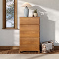 Natural wood dresser in minimalist bedroom with ceramic table lamp, potted plant and woven wicker basket beside window overlooking snowy pine trees