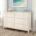 Whitewashed six-drawer wooden dresser in coastal-style bedroom with beach artwork, decorative candles, and light wood flooring
