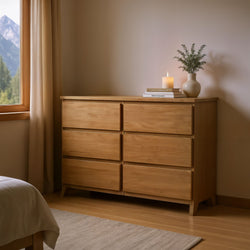 Wooden bedroom dresser with drawers, topped with books, a candle, and a vase with greenery, next to a window with mountain view.