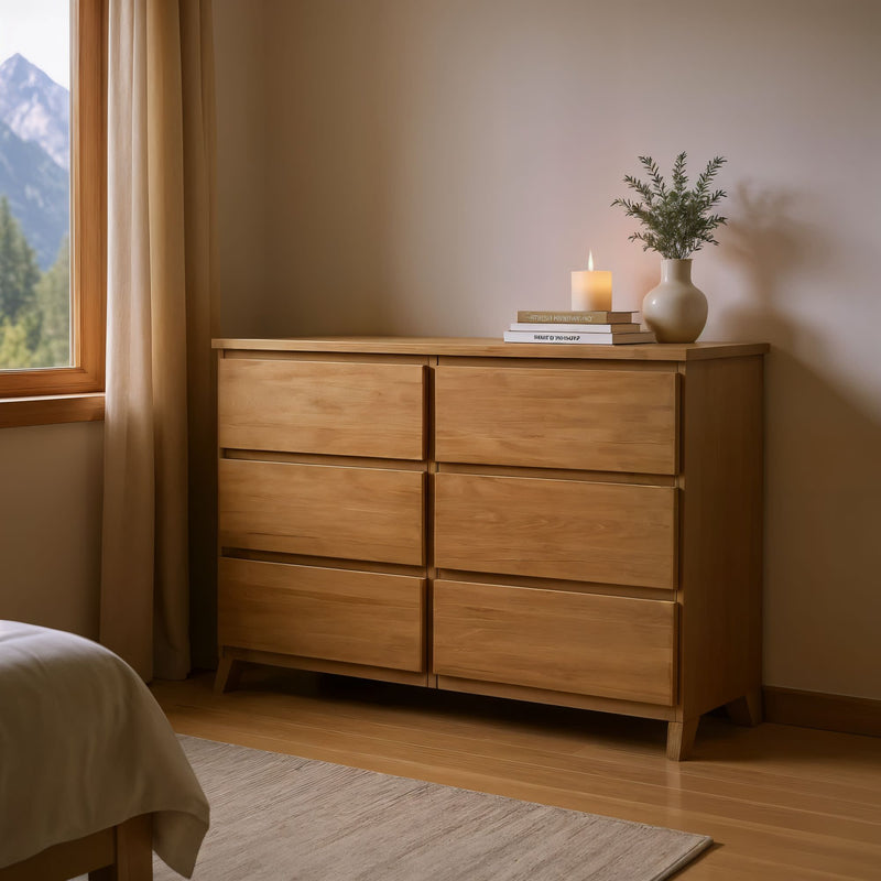 Wooden bedroom dresser with drawers, topped with books, a candle, and a vase with greenery, next to a window with mountain view.
