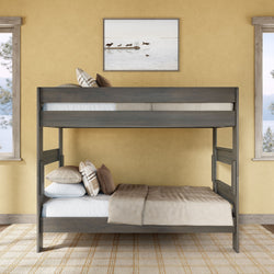 Gray wooden bunk bed in a cozy yellow bedroom with beige bedding and patterned throw pillows, framed lakeside wall art above, two windows with pine tree views and a neutral plaid area rug