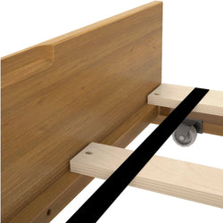 Close-up of wooden bed frame assembly with oak-finish headboard, pine slats, black central support beam and caster wheel.