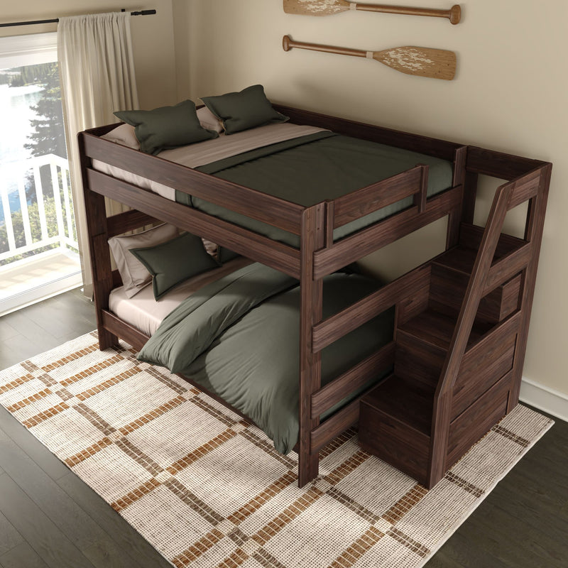 Dark walnut twin-over-full wooden bunk bed with built-in storage stairs, green bedding and pillows on a patterned rug in a bright lakeside bedroom with sliding glass door balcony