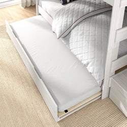 Underbed trundle mattress pulled out from whitewashed light wood bunk bed with quilted white duvet on beige rug — space-saving storage for kids' bedroom.