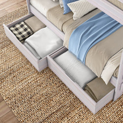 Light wood bed with pull-out underbed storage drawers filled with folded blankets, pillows and a blue throw on woven jute rug