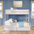 Whitewashed wooden twin-over-full bunk bed with under-bed storage drawers in a coastal blue bedroom, woven jute rug, layered neutral bedding, decorative blue pillows and light-blue throw, framed seaside artwork and ocean-view windows