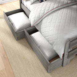 Gray wooden twin bed with built-in under-bed storage drawers holding pillows, white quilted comforter with red trim on a beige rug and hardwood floor — space-saving bedroom storage solution