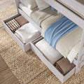 White wooden bunk bed with pull-out under-bed storage drawers holding neatly folded blankets, pillows, and a light blue throw on woven rug