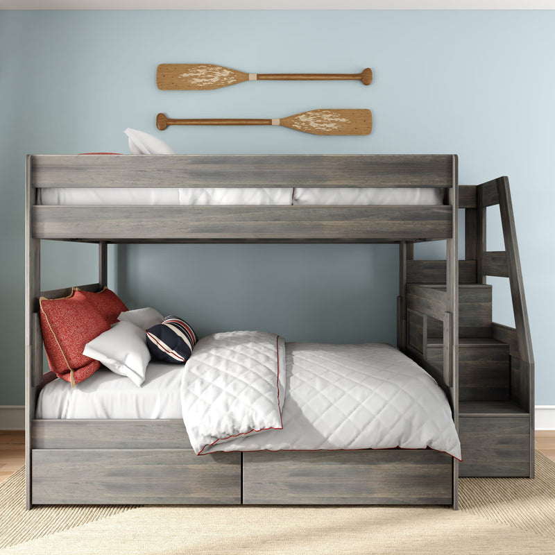 Gray wooden bunk bed with staircase and under-bed storage drawers, full lower bed and twin upper, white quilted bedding and red accent pillows against light-blue wall with wooden oars