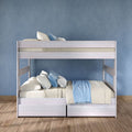 White wooden bunk bed with under-bed storage drawers, beige and blue bedding and pillows against a blue textured wall on hardwood floor