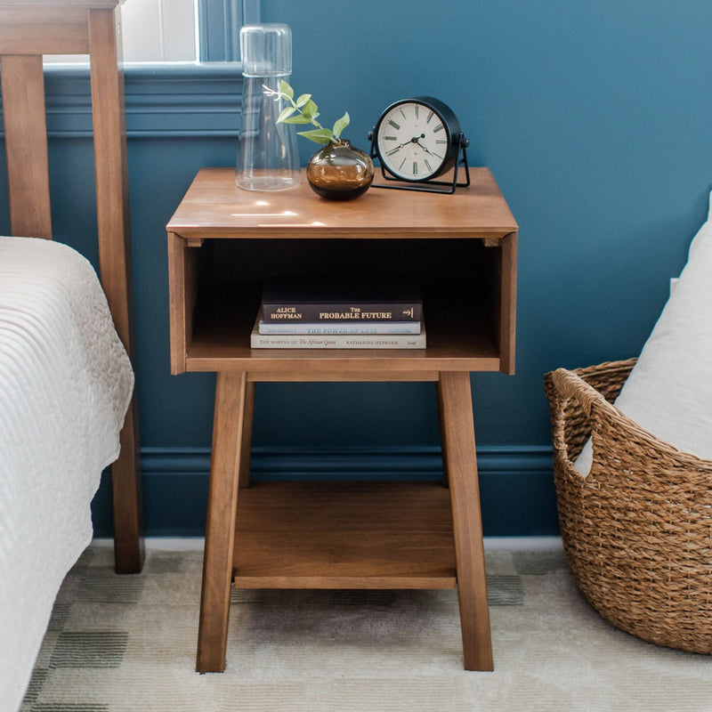 Wooden nightstand with shelf holding books and clock, glass carafe, beside woven basket and bed in modern bedroom.