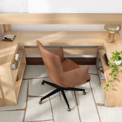 Aerial view of a modern home office with a light wood built-in desk and shelves, brown leather swivel chair on black metal casters, brass desk lamp, trailing potted plant and neutral geometric rug