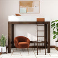 Dark walnut loft bed with ladder and white guardrail over a cozy reading nook featuring a rust velvet accent chair, round wood side table, indoor plants, rug and abstract wall art.