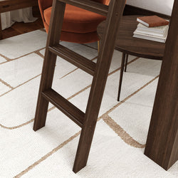 Close-up of dark walnut ladder leg in a mid-century modern living room on a white geometric area rug with a rust orange accent chair and round wooden side table with stacked books