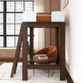 Modern dark-wood loft bed with ladder and under-bed lounge featuring an orange accent armchair, round coffee table with books, floor-to-ceiling grid windows and exposed brick wall — industrial bedroom furniture and space-saving small-space design