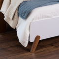 Close-up of modern white platform bed corner with angled wooden legs, white textured bedding and blue quilt on dark hardwood floor