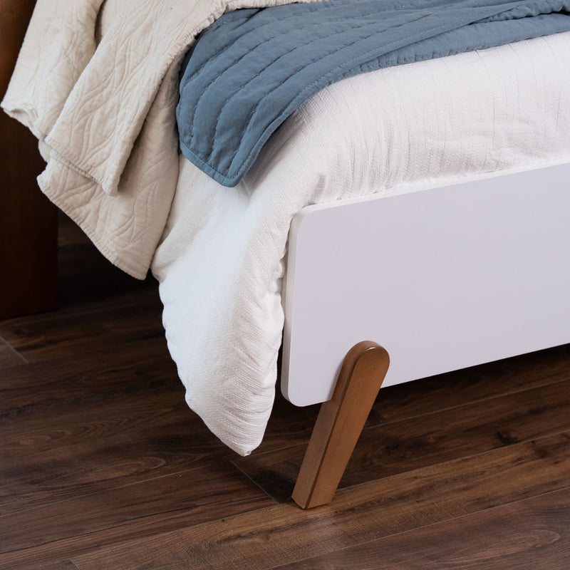 Close-up of modern white platform bed corner with angled wooden legs, white textured bedding and blue quilt on dark hardwood floor