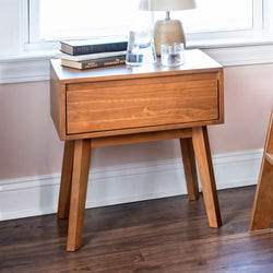 Oak-finish mid-century modern wooden nightstand with single drawer, stacked books and lamp on hardwood floor by window