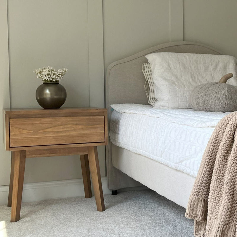 Cozy bedroom interior with beige tones featuring a wooden nightstand, decorative vase with flowers, and a stylish bed with white bedding and a knitted throw blanket.