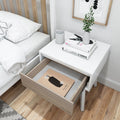 Modern white bedside table with an open drawer showcasing a book, stacked books on top, and a small vase with greenery, in a minimalist bedroom setting.