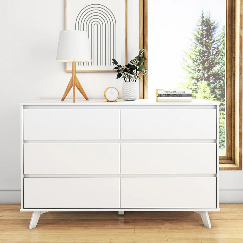 White modern dresser with six drawers, featuring a wooden lamp, decorative clock, and plants, set beside a window with a nature view.