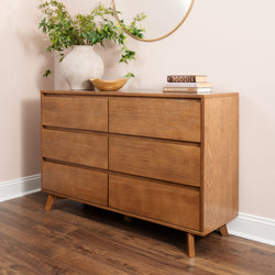 Mid-century modern six-drawer wooden dresser in natural oak finish on hardwood floor, styled with a ceramic vase of greenery, wooden bowl, stacked books and a round wall mirror in a neutral bedroom.