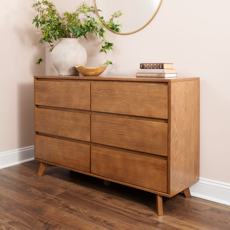 Mid-century modern six-drawer wooden dresser in natural oak finish on hardwood floor, styled with a ceramic vase of greenery, wooden bowl, stacked books and a round wall mirror in a neutral bedroom.