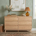 Light wood mid-century dresser against sage green wall with pampas grass vase, amber glass lamp, stacked books and neutral decor — modern minimalist bedroom storage and styling