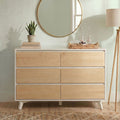 Minimalist light wood dresser with round gold mirror, snake plant on stacked books, wicker tray with glass bottles and bowl, tripod floor lamp in a modern entryway
