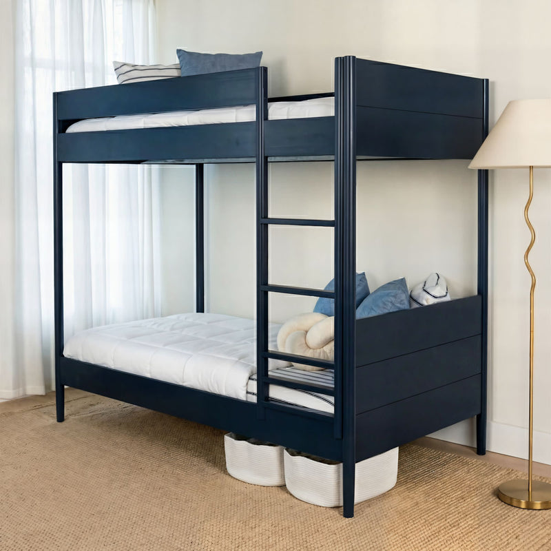 Navy blue wooden bunk bed with ladder in a modern bedroom, white bedding and blue accent pillows, floor lamp and under-bed storage baskets