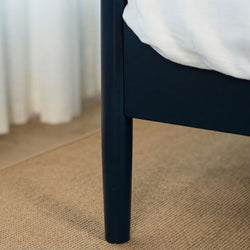 Close-up of a black bed frame leg on a natural jute rug with white bedding and sheer curtains, minimalist bedroom decor