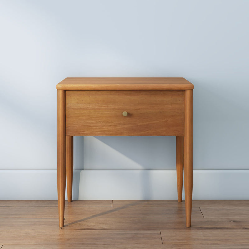 Mid-century modern oak bedside nightstand with single drawer and brass knob, tapered legs, light wood finish against pale blue wall on hardwood floor