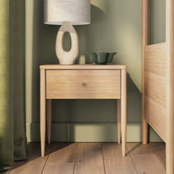 Scandinavian oak bedside table with single drawer, sculptural table lamp and green ceramic bowl on hardwood floor against a sage green bedroom wall