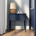 Navy blue bedside table with woven rattan lamp, lit scented candle on marble tray, hardwood floor and white curtains in a modern bedroom