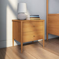 Mid-century modern two-drawer oak bedside table with tapered legs, white textured table lamp and stacked books in a bright minimalist bedroom