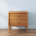 Mid-century modern light wood two-drawer bedside table with brass knobs on hardwood floor against pale blue wall