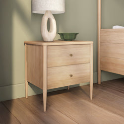 Scandinavian light oak two-drawer nightstand with tapered legs, table lamp and green decorative bowl on hardwood bedroom floor against a sage green wall