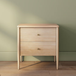 Light oak two-drawer bedside table nightstand with brass knobs against sage green wall on hardwood floor, minimalist mid-century modern bedroom furniture