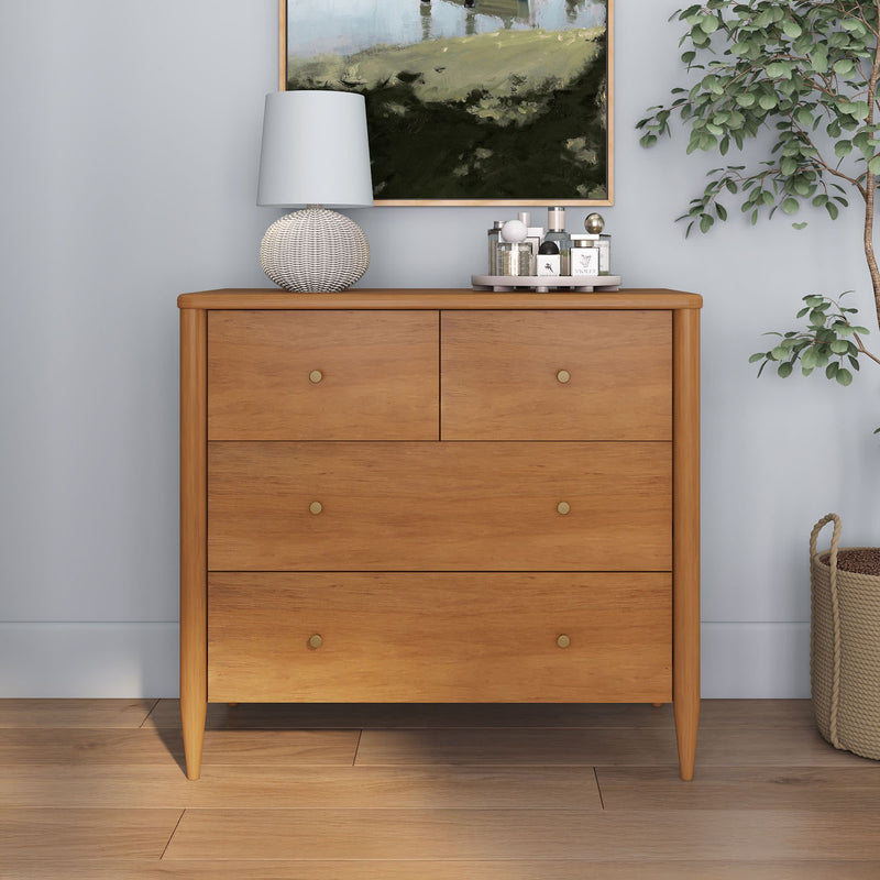 Mid-century modern light wood dresser with four drawers in a styled bedroom, woven table lamp, perfume tray, framed landscape artwork and potted plant