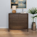 Dark walnut four-drawer dresser with brass knobs in a coastal living room vignette, topped with a woven table lamp, reed diffuser and framed sailboat painting, beside a potted palm on hardwood floor